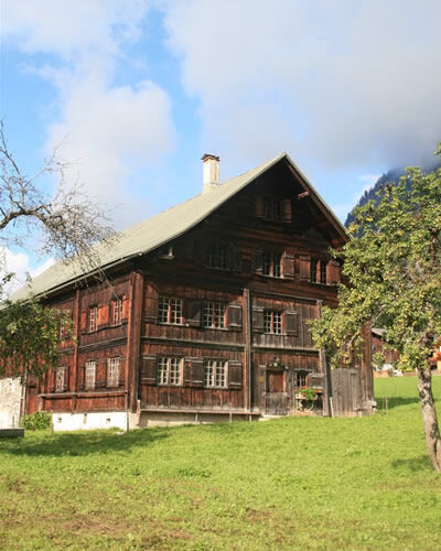 Ein großes Holzhaus mit mehreren Fenstern steht auf einem grasbewachsenen Rasen unter einem teilweise bewölkten Himmel, in dessen Nähe sich ein paar Bäume befinden.