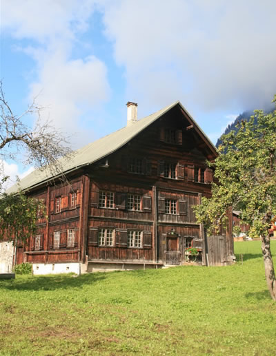 Ein großes Holzhaus mit mehreren Fenstern steht auf einem grasbewachsenen Rasen unter einem teilweise bewölkten Himmel, in dessen Nähe sich ein paar Bäume befinden.