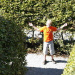 Ein Junge in einem orangefarbenen Hemd und grauen Shorts steht mit erhobenen Armen auf einem Kiesweg zwischen grünen Hecken.