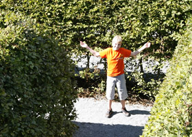 Ein Junge in einem orangefarbenen Hemd und grauen Shorts steht mit erhobenen Armen auf einem Kiesweg zwischen grünen Hecken.