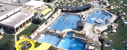 Luftaufnahme eines öffentlichen Freibadkomplexes mit Wasserrutschen, mehreren Becken, Sonnenliegen und grünen Rasenflächen.