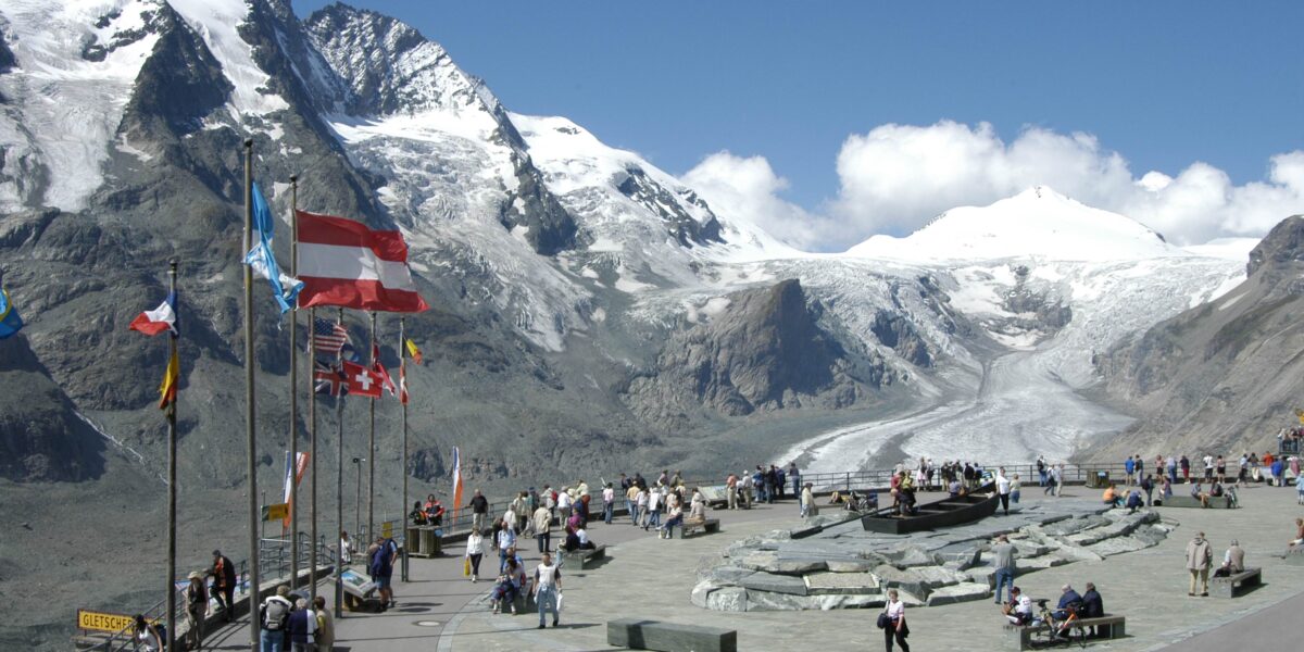 Die Touristen versammeln sich auf einer Aussichtsplattform mit mehreren Fahnen und blicken auf schneebedeckte Berge und einen Gletscher unter einem klaren blauen Himmel.