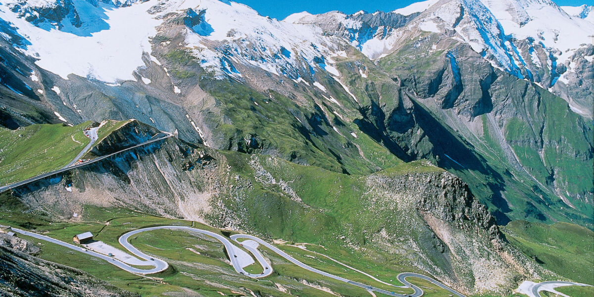 Eine kurvenreiche Bergstraße mit mehreren scharfen Kurven, umgeben von grünen Hängen und schneebedeckten Gipfeln unter strahlend blauem Himmel.