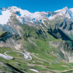 Eine kurvenreiche Bergstraße mit mehreren scharfen Kurven, umgeben von grünen Hängen und schneebedeckten Gipfeln unter strahlend blauem Himmel.