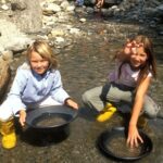 Zwei Kinder knien in einem flachen, felsigen Bach und halten jeweils eine schwarze Pfanne ins Wasser, wahrscheinlich um nach Gold zu wühlen.
