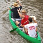 Drei Personen in einem grünen Kanu paddeln einen Fluss hinunter; zwei tragen Strohhüte und eine ein weißes Hemd mit roter Aufschrift.