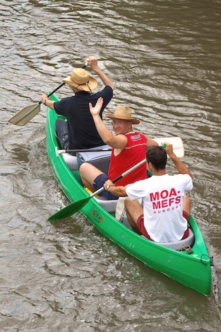 Drei Personen in einem grünen Kanu paddeln einen Fluss hinunter; zwei tragen Strohhüte und eine ein weißes Hemd mit roter Aufschrift.