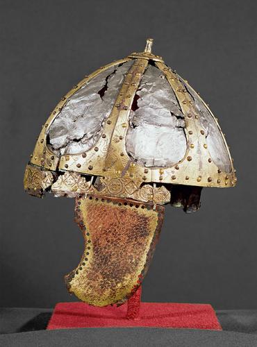 Ein korrodierter antiker Helm mit kuppelförmigem Metallrahmen, Ziernieten und beschädigtem Wangenschutz, ausgestellt auf einem roten Ständer vor einem schlichten Hintergrund.