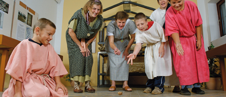 Kinder und Erwachsene in historischen Kostümen spielen drinnen ein traditionelles Spiel und beobachten, wie ein Gegenstand auf dem Boden rollt.