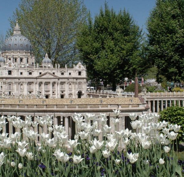 Eine detailgetreue Miniaturnachbildung des Petersdoms steht umgeben von Bäumen und blühenden weißen Tulpen in einer Parklandschaft.