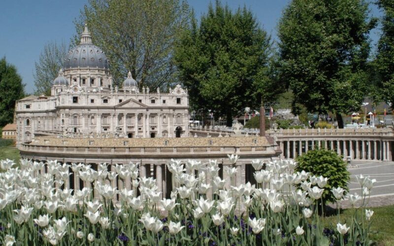 Eine detailgetreue Miniaturnachbildung des Petersdoms steht umgeben von Bäumen und blühenden weißen Tulpen in einer Parklandschaft.