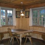 Eine hölzerne Essecke mit einem sechseckigen Tisch, zwei Stühlen, einer Sitzbank, zwei Fenstern mit weißen Vorhängen und zwei gerahmten Bildern an den getäfelten Wänden.