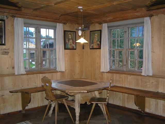Eine hölzerne Essecke mit einem sechseckigen Tisch, zwei Stühlen, einer Sitzbank, zwei Fenstern mit weißen Vorhängen und zwei gerahmten Bildern an den getäfelten Wänden.