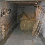 In einem schmalen Raum mit Steinwänden sind Holzschlitten, Heuballen, alte landwirtschaftliche Geräte und Schwarz-Weiß-Fotos an der linken Wand ausgestellt.
