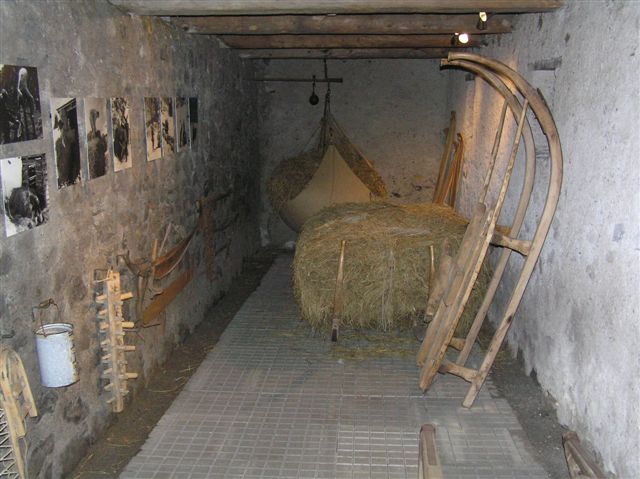 In einem schmalen Raum mit Steinwänden sind Holzschlitten, Heuballen, alte landwirtschaftliche Geräte und Schwarz-Weiß-Fotos an der linken Wand ausgestellt.
