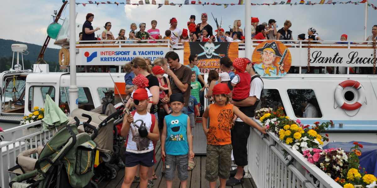 Eine Gruppe von Kindern und Erwachsenen, viele von ihnen in Piratenkostümen, steht auf einem mit Fahnen und Blumen geschmückten Boot im Hafen.