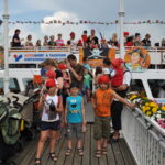 Eine Gruppe von Kindern und Erwachsenen, viele von ihnen in Piratenkostümen, steht auf einem mit Fahnen und Blumen geschmückten Boot im Hafen.