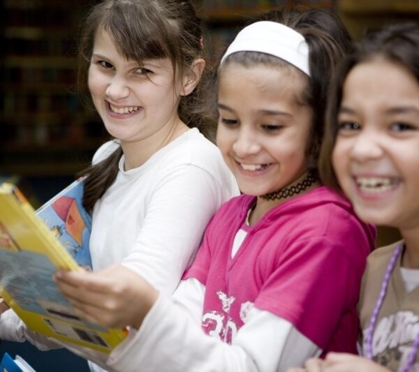 Drei Mädchen stehen zusammen, lächeln und lachen, während sie sich in einer Bibliothek Bücher ansehen.