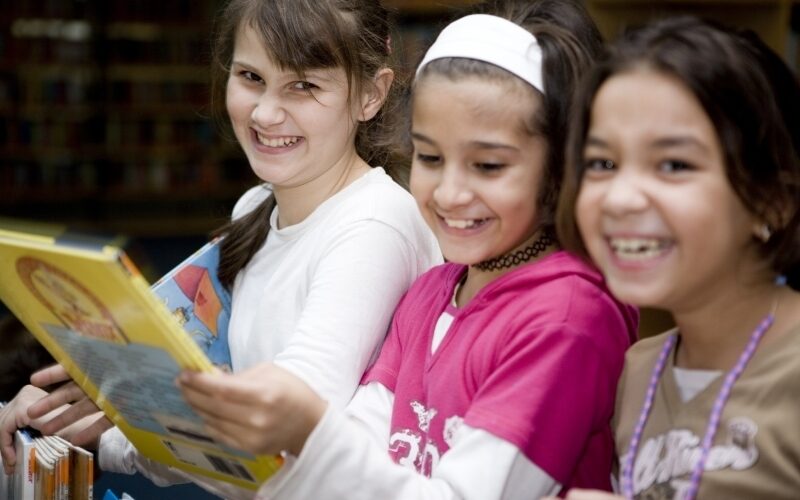 Drei Mädchen stehen zusammen, lächeln und lachen, während sie sich in einer Bibliothek Bücher ansehen.