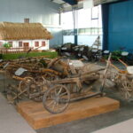 In den Innenräumen sind auf Podesten mehrere alte hölzerne landwirtschaftliche Geräte und Pflüge ausgestellt, mit einem Modell eines strohgedeckten Bauernhauses im Hintergrund.