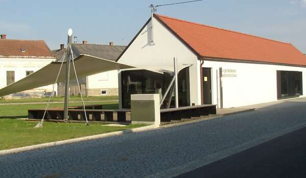 Ein modernes, einstöckiges Gebäude mit rotem Ziegeldach und weißer Fassade, das einen großen dreieckigen Sonnenschirm über einer kleinen Terrasse aufweist.