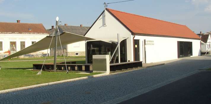 Ein modernes, einstöckiges Gebäude mit rotem Ziegeldach und weißer Fassade, das einen großen dreieckigen Sonnenschirm über einer kleinen Terrasse aufweist.