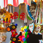 An einem bunten Marktstand werden unter einem gestreiften Vordach verzierte Lebkuchenherzen mit deutschen Texten, Spielzeugautos und Scherzbrillen angeboten.