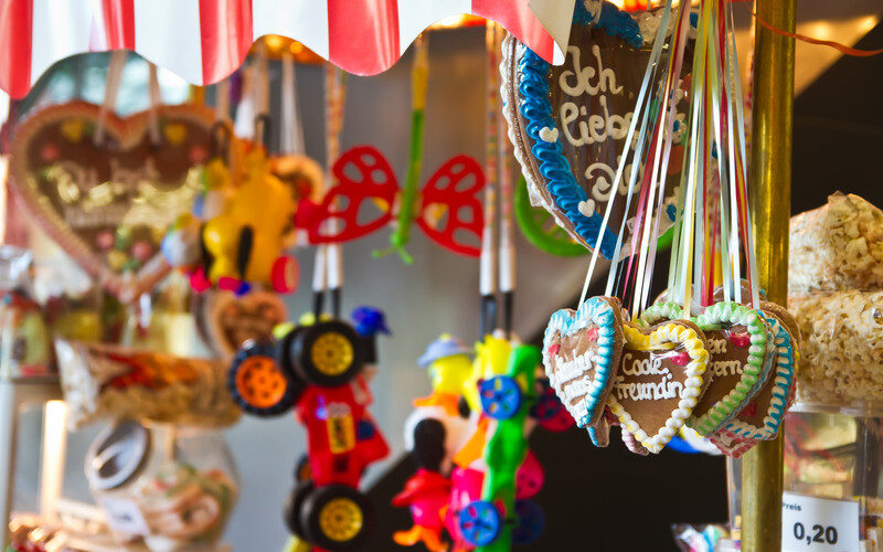 An einem bunten Marktstand werden unter einem gestreiften Vordach verzierte Lebkuchenherzen mit deutschen Texten, Spielzeugautos und Scherzbrillen angeboten.