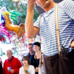 Ein Clown im gestreiften Hemd und mit Hosenträgern hält eine Trompete und eine rote Nase in der Hand und tritt bei einer Indoor-Veranstaltung vor einer Menge von Erwachsenen und Kindern auf.