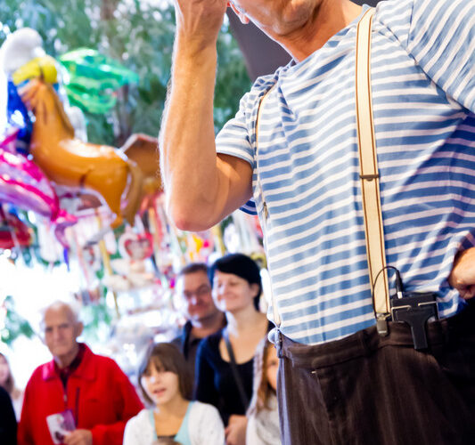 Ein Clown im gestreiften Hemd und mit Hosenträgern hält eine Trompete und eine rote Nase in der Hand und tritt bei einer Indoor-Veranstaltung vor einer Menge von Erwachsenen und Kindern auf.