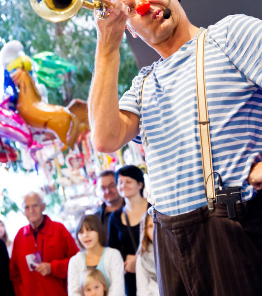 Ein Clown im gestreiften Hemd und mit Hosenträgern hält eine Trompete und eine rote Nase in der Hand und tritt bei einer Indoor-Veranstaltung vor einer Menge von Erwachsenen und Kindern auf.