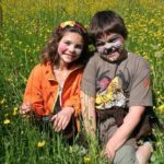 Zwei Kinder mit bemalten Gesichtern sitzen an einem sonnigen Tag dicht beieinander in einem Feld mit hohem grünen Gras und gelben Wildblumen.
