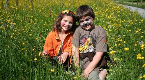 Zwei Kinder mit bemalten Gesichtern sitzen an einem sonnigen Tag dicht beieinander in einem Feld mit hohem grünen Gras und gelben Wildblumen.