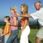 Vier Personen und ein Kind stehen in einer Reihe im Freien auf einer Wiese, jeder hat die Hände auf den Schultern des Vordermanns, im Hintergrund ist eine malerische Landschaft zu sehen.