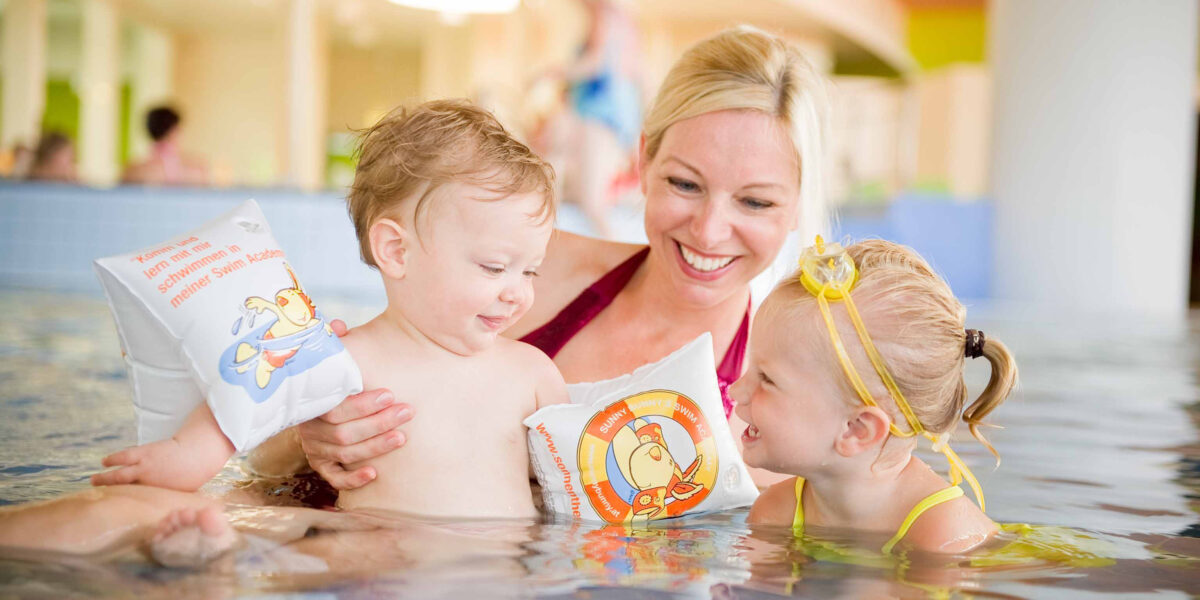 Ein Erwachsener und zwei kleine Kinder mit Schwimmarmbändern lächeln und spielen zusammen in einem Schwimmbad.