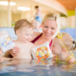 Ein Erwachsener und zwei kleine Kinder mit Schwimmarmbändern lächeln und spielen zusammen in einem Schwimmbad.