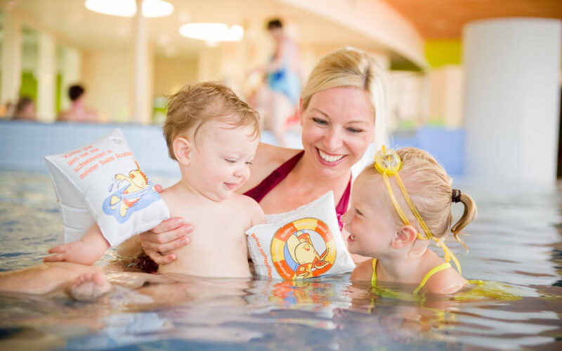 Ein Erwachsener und zwei kleine Kinder mit Schwimmarmbändern lächeln und spielen zusammen in einem Schwimmbad.