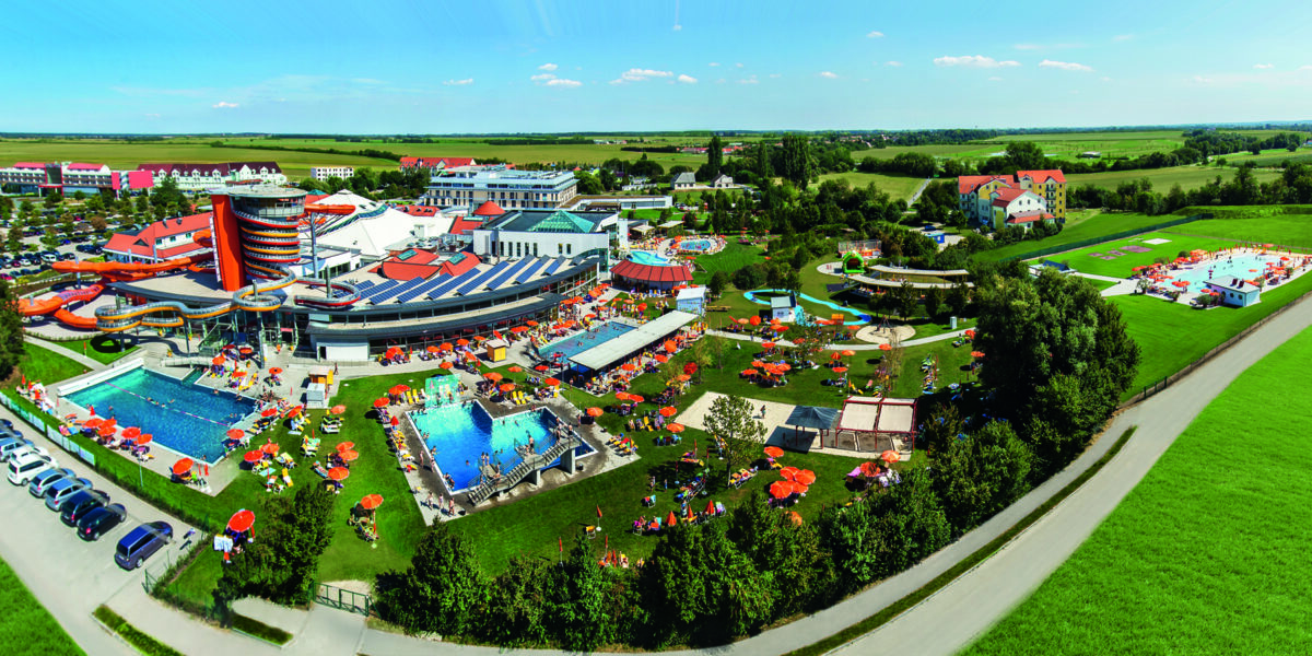 Luftaufnahme eines großen Wasserparks im Freien mit Schwimmbecken, Wasserrutschen, Liegestühlen, Sonnenschirmen und umliegenden grünen Feldern unter einem klaren Himmel.