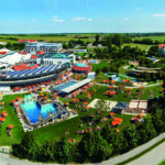 Luftaufnahme eines großen Wasserparks im Freien mit Schwimmbecken, Wasserrutschen, Liegestühlen, Sonnenschirmen und umliegenden grünen Feldern unter einem klaren Himmel.