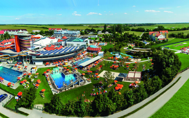 Luftaufnahme eines großen Wasserparks im Freien mit Schwimmbecken, Wasserrutschen, Liegestühlen, Sonnenschirmen und umliegenden grünen Feldern unter einem klaren Himmel.