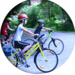 Drei Kinder mit Helmen fahren mit dem Fahrrad auf einem gepflasterten Weg in einem bewaldeten Außenbereich.