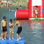 Kinder und Erwachsene schwimmen und spielen an einem sonnigen Tag auf schwimmenden Plattformen und Schlauchbooten in einem See.