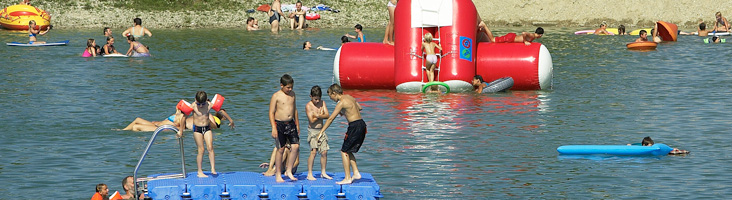 Kinder und Erwachsene schwimmen und spielen an einem sonnigen Tag auf schwimmenden Plattformen und Schlauchbooten in einem See.