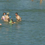 Menschen schwimmen und spielen mit einem Ball in einem ruhigen Gewässer in der Nähe eines Kiesstrandes an einem sonnigen Tag.