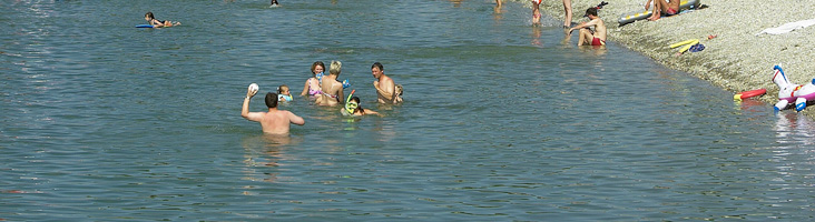 Menschen schwimmen und spielen mit einem Ball in einem ruhigen Gewässer in der Nähe eines Kiesstrandes an einem sonnigen Tag.