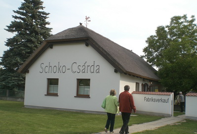 Zwei Personen gehen auf ein kleines weißes Gebäude mit einem Strohdach zu, das von außen mit "Schoko-Csárda" und "Fabrikverkauf" beschriftet ist.