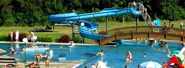 Außenschwimmbad mit schwimmenden, faulenzenden und spielenden Menschen; eine Wasserrutsche und eine Holzbrücke führen über das Becken; im Hintergrund eine Wiese und Bäume.