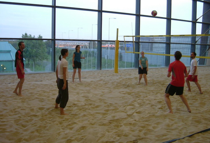 Sechs Personen spielen Volleyball auf einem überdachten Sandplatz mit großen Fenstern im Hintergrund.