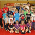 Eine Gruppe von Kindern und Erwachsenen posiert für ein Foto in einer Sporthalle, einige halten Tischtennisschläger, im Hintergrund sind Tischtennisplatten zu sehen.