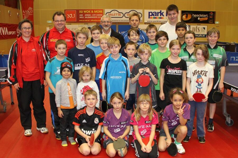 Eine Gruppe von Kindern und Erwachsenen posiert für ein Foto in einer Sporthalle, einige halten Tischtennisschläger, im Hintergrund sind Tischtennisplatten zu sehen.